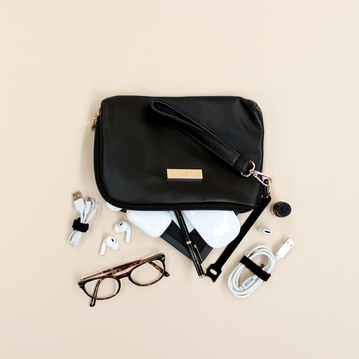Black faux leather cable organiser bag with zip, removable strap, tech loops and multiple storage pockets. Filled with air pods, mouse, charging cables and glasses.